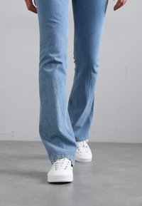 Lichtblauwe flare jeans met een vervaagde textuur, gecombineerd met witte sneakers, weergegeven tegen een neutrale grijze achtergrond.