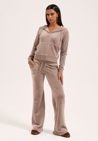 Lichtbeige, fluwelen trainingspak met een rits hoodie en broek met elastische taille. Bevat voorzakken en heeft een comfortabele pasvorm. Platte schoenen maken de look af.