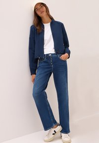 Navy crop-jakke med elastiske mansjetter, hvit T-skjorte, høytlivede blå jeans og hvite sneakers med svarte detaljer.