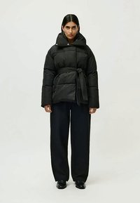 Femme portant une doudoune noire ceinturée, un pantalon noir à large jambe et des chaussures noires, se tenant contre un fond gris clair.