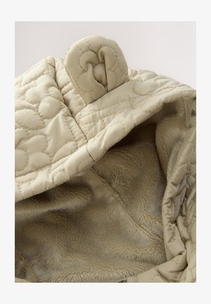 Beżowa pikowana tkanina z miękką, sztuczną podszewką futerkową. Wnętrze ma gładką teksturę i pętelkę do zamknięcia.