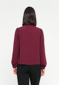 Blouse bordeaux à manches longues en tissu texturé, avec des manches ajustées et un col arrondi. Portée sur un pantalon noir, elle affiche une coupe décontractée.