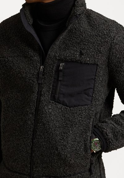 Strukturirana črna flis jakna z žepom na prsih z zadrgo in gladkim obrobo. Ima zadrgo za zapiranje in pritrjen detajl za uro.