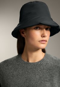 Pălărie neagră dintr-un material neted, cu un design lat și cu marginea coborâtă. Pulover gri tricotat cu un gât rotund.