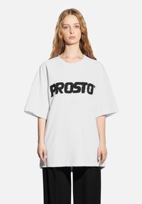 Biała, oversize'owa koszulka z krótkim rękawem, z wyraźnym czarnym napisem "PROSTO" na froncie. Wykonana z bawełny o gładkiej fakturze.