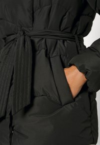 Cappotto imbottito nero con vita cinturata, caratterizzato da una texture liscia, design trapuntato e tasche laterali con dettaglio arricciato.