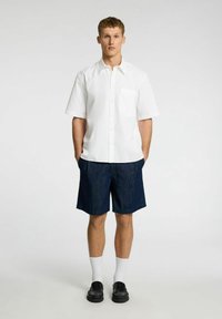 Camicia bianca a maniche corte con taschino, abbinata a pantaloni corti in denim scuro. Il modello indossa calze bianche e scarpe nere. Outfit semplice e casual.