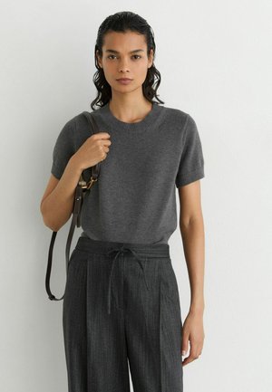 Donna con capelli scuri e bagnati indossa un maglione grigio a maniche corte e pantaloni larghi grigio scuro a righe sottili, tenendo la tracolla di una borsa scura.