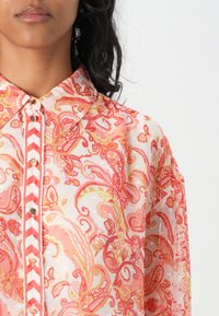 Sheer, long-sleeve blouse featuring a floral and paisley pattern in pink, orange, and yellow, with a button-up front and chevron accents.