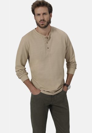 Beige Langarm-Henley-Shirt mit drei vorderen Knöpfen, aus weichem Stoff, kombiniert mit olivgrünen Hosen. Das Modell steht mit den Händen in den Taschen.