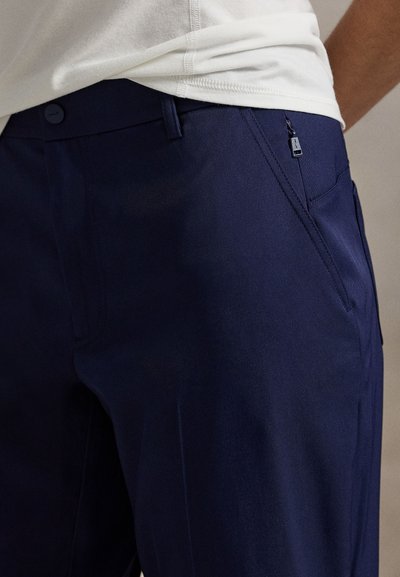 Tumši zili bikses no gludas auduma ar šauru piegriezumu, kas aprīkotas ar priekšējām kabatām un nelielu rāvējslēdzēja detaļu sānos.