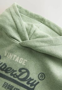 Sudadera verde claro con capucha, con un tejido texturizado y la marca "Vintage SuperDry" en texto oscuro en la parte delantera.