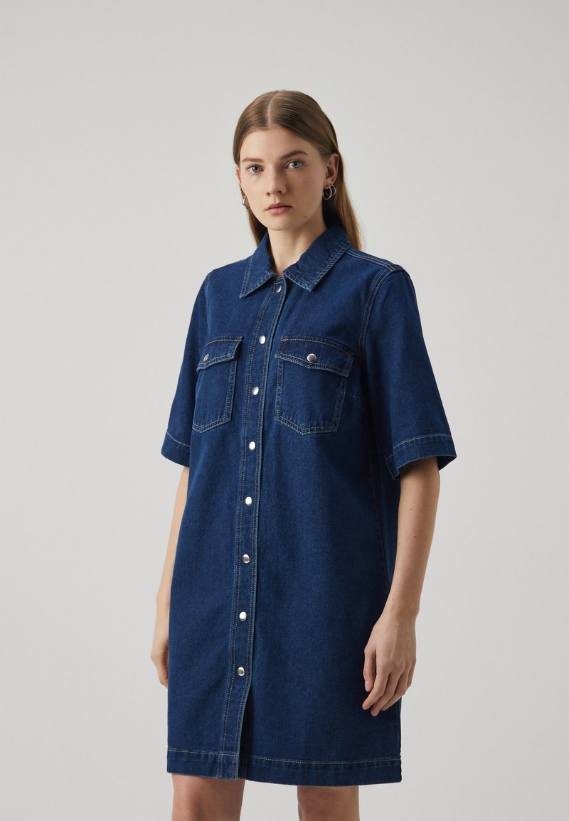 Robe chemise en denim bleu foncé avec des manches courtes, deux poches poitrine, une fermeture à boutons sur le devant et des détails de couture. Texture lisse.
