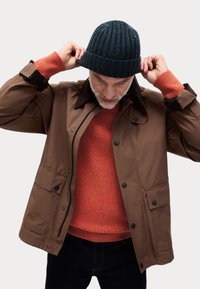 Chaqueta marrón impermeable con botones negros, cuello marrón oscuro, llevada sobre un suéter naranja texturizado y un gorro azul de canalé.