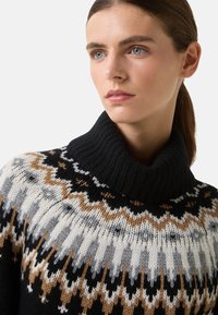 Vrouw met blauwe ogen die een zwarte coltrui draagt met witte, bruine en grijze geometrische breipatronen, die iets naar rechts kijkt.