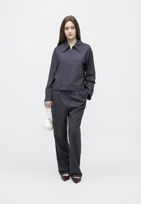 Mladá žena s dlhými hnedými vlasmi, oblečená v tmavosivej bunde na zips a zodpovedajúcich nohaviciach, držiaca malú bielu kabelku, stojaca na bielom pozadí.