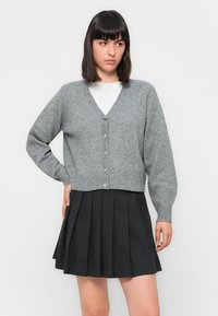 Frau mit glattem, dunklem Haar, die eine graue, knöpfbare Strickjacke über einem weißen Oberteil und einem schwarzen, gefalteten Rock trägt und vor einem weißen Hintergrund steht.