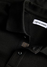 Polo noir côtelé avec col, bouton et étiquette visible de la marque Jack & Jones à l'intérieur de l'encolure.