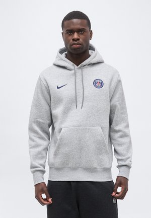 Mladý muž čelom vpred, oblečený v svetlosivej mikine s kapucňou Paris Saint-Germain s logom Nike a predným vreckom, stojaci pred jednofarebným pozadím.