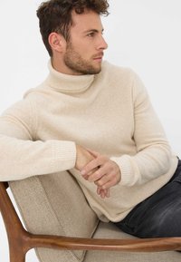 Maglione beige a dolcevita in maglia morbida e strutturata. Design aderente con polsini a coste. Modella seduta su una sedia imbottita beige con bracciolo in legno.