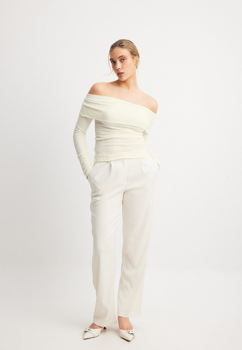 Cremefarbenes, strukturiertes Oberteil mit schulterfreiem Design, figurbetonter Taille; kombiniert mit hochgeschnittenen cremfarbenen Hosen und weißen Schuhen mit spitzem Zehenbereich und Akzenten.
