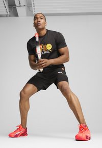 Schwarzes Sportshirt mit orangefarbigem Grafik, schwarze Shorts und knallrote Sportschuhe. Hält einen Padel-Schläger und ist in einer dynamischen Pose.