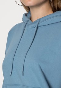 Felpa blu con cappuccio e coulisse, realizzata in tessuto morbido, con tasca frontale e texture liscia. Dettagli di cuciture resistenti a completare il design.