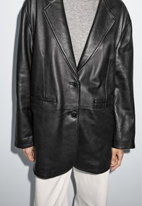 Černý kožený blazer s klopovým límcem, zapínáním na jeden knoflík a bočními kapsami. Nošený přes šedý top, vyznačuje se hladkou texturou.