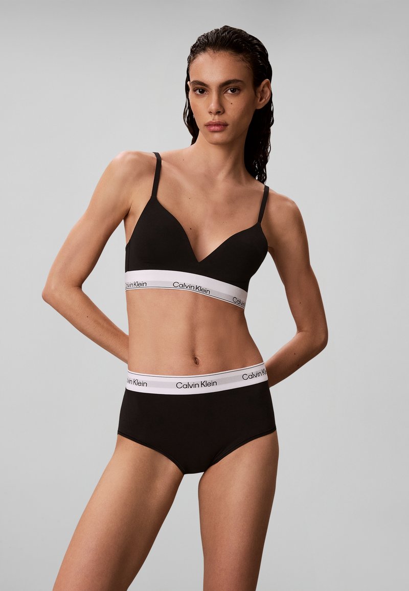 Γυναίκα φοράει μαύρο σουτιέν και εσώρουχο Calvin Klein με λευκές ελαστικές λωρίδες με λογότυπο σε μονόχρωμο γκρι φόντο.