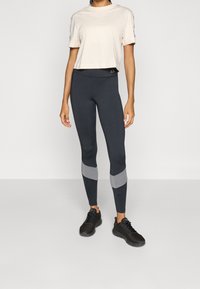 Kobieta ubrana w krótką białą koszulkę, ciemnoszare legginsy z jasnoszarymi pasami poniżej kolan oraz czarne buty sportowe, stojąca na gładkiej podłodze.