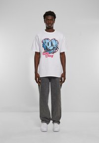 Hvid t-shirt med korte ærmer og et blåt grafisk design med et smilende ansigt og teksten "sad boy" i pink. Båret med grå denimbukser og hvide sko.