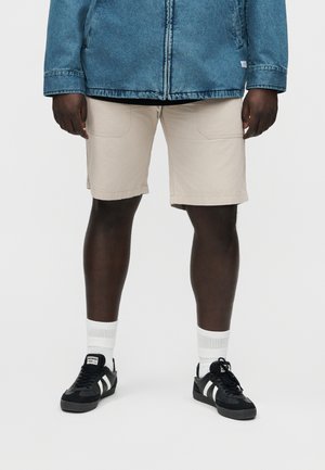 Tamsios odos žmogus dėvi smėlio spalvos šortus, baltas kojines, juodos ir baltos spalvų sportbačius bei mėlyną džinsinį švarką, stovintis priešais paprastą foną.