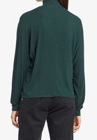 Maglietta a maniche lunghe verde scuro con colletto alto, texture a coste e spacchi laterali, abbinata a pantaloni in denim neri. Design semplice, silhouette aderente.