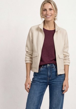 Smilende kvinde iført en cremefarvet lynlåstrøje over en bordeaux skjorte og blå jeans, stående mod en ensartet lys baggrund.