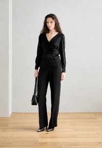 Top nero avvolgente con tessuto texture, scollatura a V profonda e maniche lunghe, abbinato a pantaloni neri su misura e scarpe nere a punta.