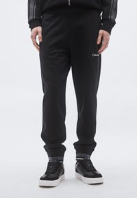 Calças de jogging pretas feitas de tecido macio, apresentando um corte afunilado, punhos elásticos e um pequeno logótipo branco "Calvin" na coxa.