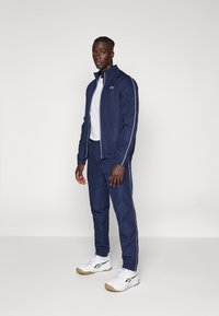 Marineblå træningsdragt lavet af et glat stof, der består af en jakke med lynlås, hvide striber og et logo, samt matchende bukser og hvide sneakers.