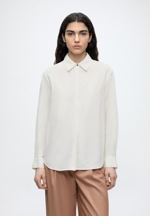 Γυναίκα με ίσια σκούρα μαλλιά, ντυμένη με άσπρο πουκάμισο με μακριά μανίκια και καφέ παντελόνι, στέκεται μπροστά σε ένα απλό φόντο.