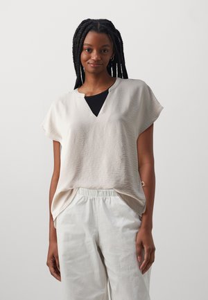 Haut beige clair à manches courtes avec un tissu texturé, présentant un accent en V noir, porté sur un pantalon blanc à cordon.
