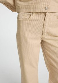 Pantalons rayés beiges en coton, avec un design classique à cinq poches, fermeture à bouton et texture subtile.