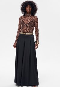 Top z długim rękawem w brązowe tonacje ze wzorem węża; w połączeniu z czarnymi szerokimi spodniami z wysokim stanem, ozdobionymi dekoracyjnymi ćwiekami na pasku.