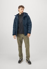 Marineblauer wasserdichter Parka mit Kapuze, Reißverschluss vorne und elastischen Bündchen, getragen über einem schwarzen Pullover, kombiniert mit olivfarbenen Cargo-Hosen und schwarzen Stiefeln.