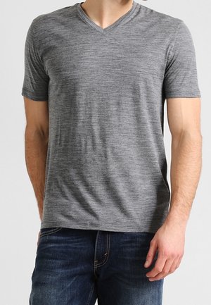 Mężczyzna ubrany w szarą koszulkę z krótkim rękawem i dekoltem w serek, zestawioną z ciemnoniebieskimi dżinsami, stojący z jedną ręką swobodnie opuszczoną wzdłuż ciała.