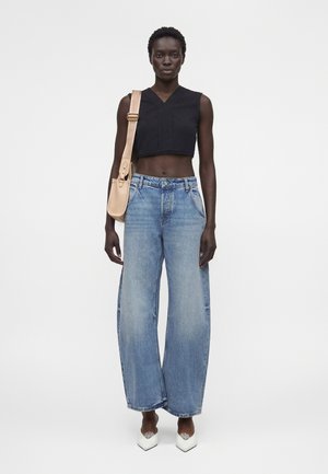 Kvinde iført en sort ærmeløs crop top, vide blå jeans, hvide spidse sko og bærende en beige skuldertaske på hvid baggrund.