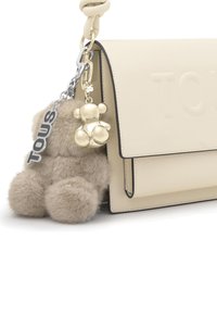 Bolso de mano de cuero crema con ribete negro y nombre de la marca grabado en relieve, adornado con un llavero de oso beige esponjoso y un charm metálico brillante en forma de osito.