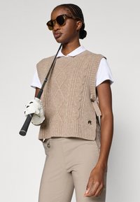 Pulover bej fără mâneci, tricotat cu model în formă de cablu, combinat cu o cămașă albă cu guler și pantaloni bej, ținând un baston de golf, purtând ochelari de soare.