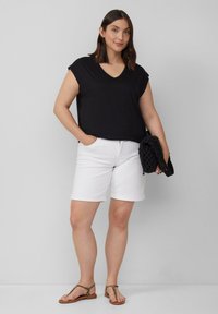 Zwarte, korte mouwen, V-hals top gecombineerd met witte denim shorts. Model houdt een zwarte, gestructureerde tas vast en draagt bruine, gespen sandalen.
