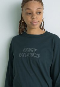 Crna majica s dugim rukavima koja sadrži tekst "OBEY STUDIOS" u svijetlosivoj boji. Tkanina izgleda mekano s opuštenim krojem.