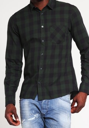 Mężczyzna ubrany w ciemnozieloną i czarną koszulę na guziki w kratę z kieszenią na piersi oraz jasnoniebieskie, podniszczone jeansy.