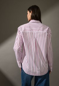 Camicia a righe bianche e rosa con piega sul retro, polsini abbottonati e colletto classico. Il materiale sembra leggero e con texture.